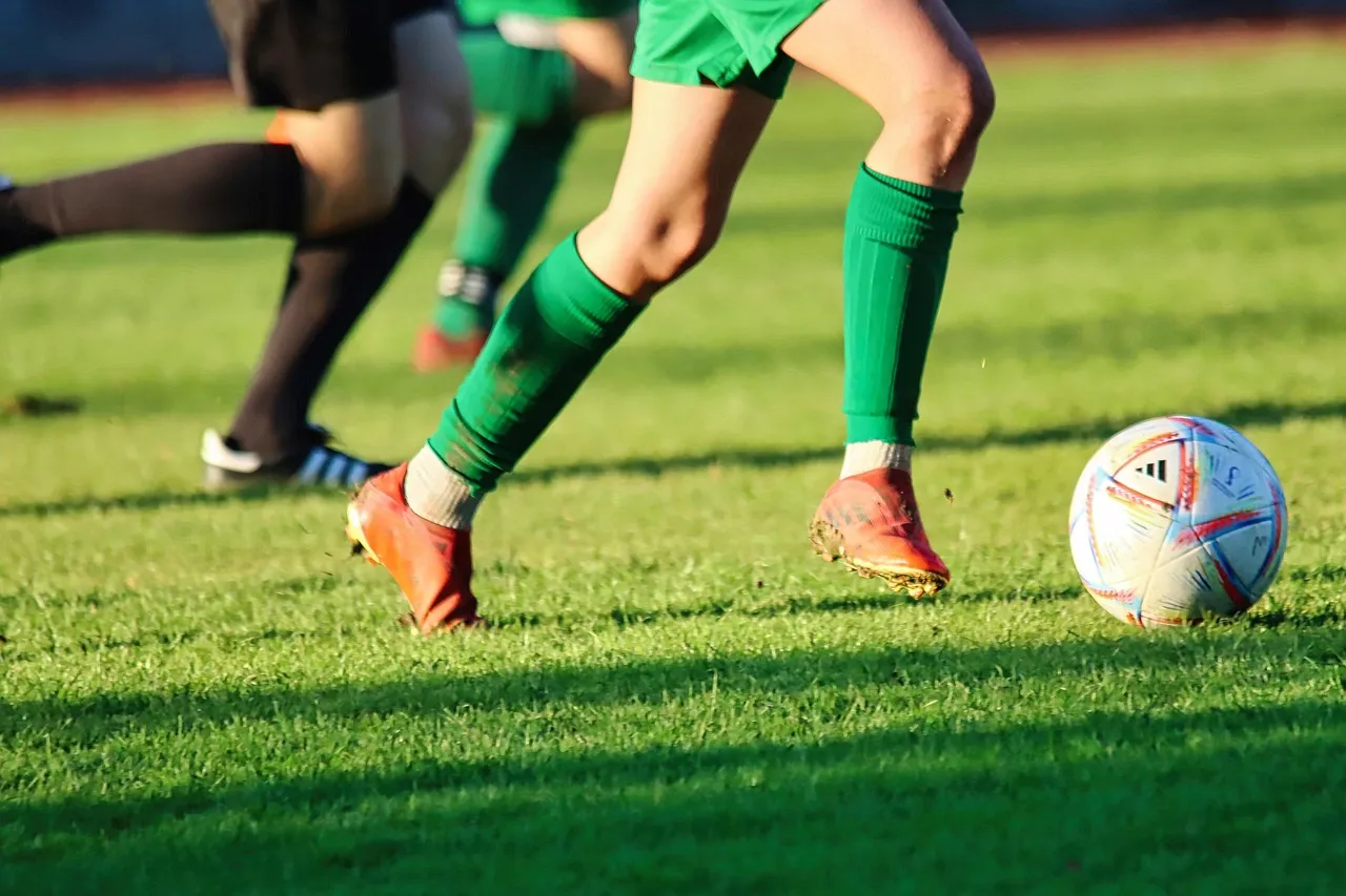 Players in green kits chasing a ball across a sunlit grass pitch