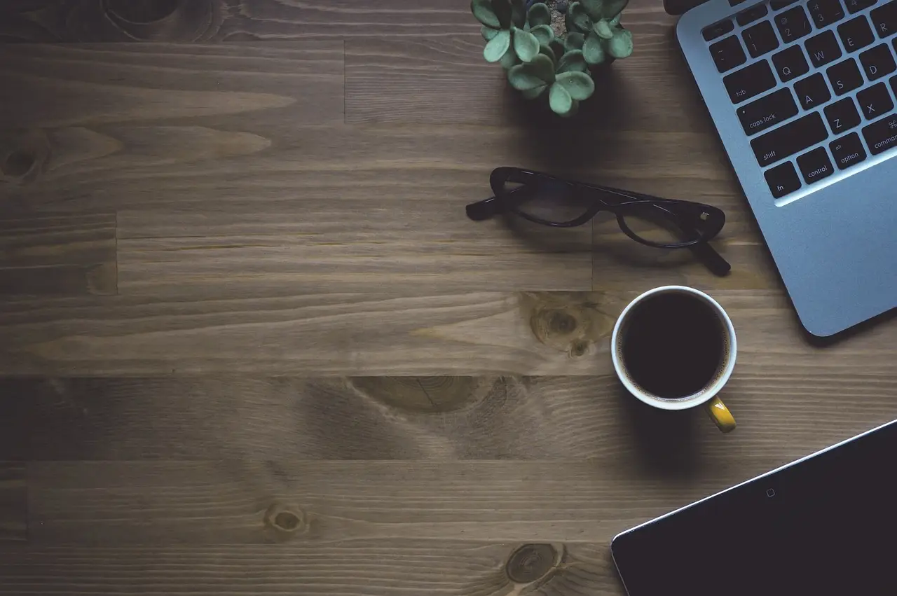 Overhead view of a wooden desk with an open laptop, black coffee, eyeglasses, tablet and a succulent