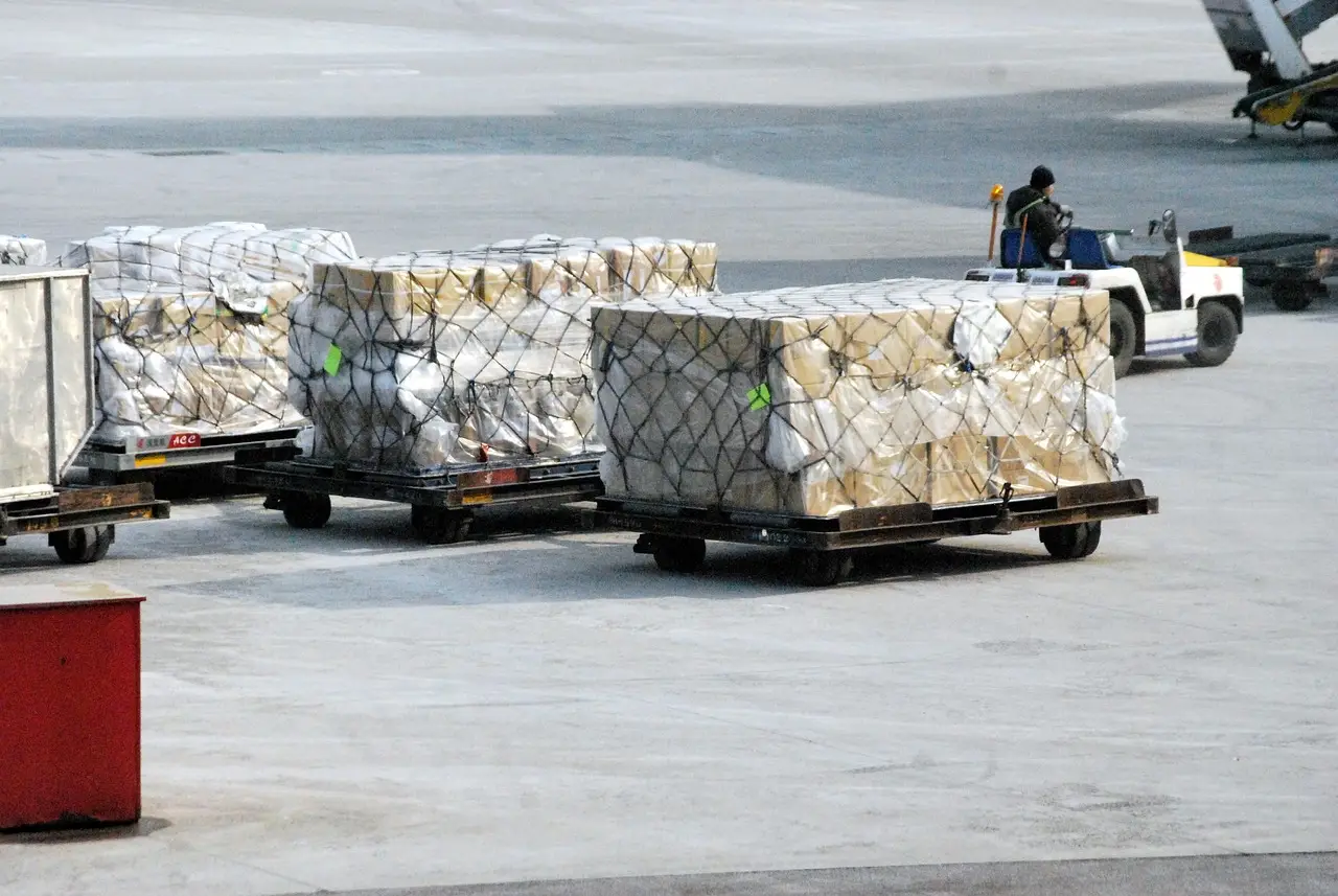 Pallets of net-wrapped freight on airport dollies being towed across the tarmac by a seated driver