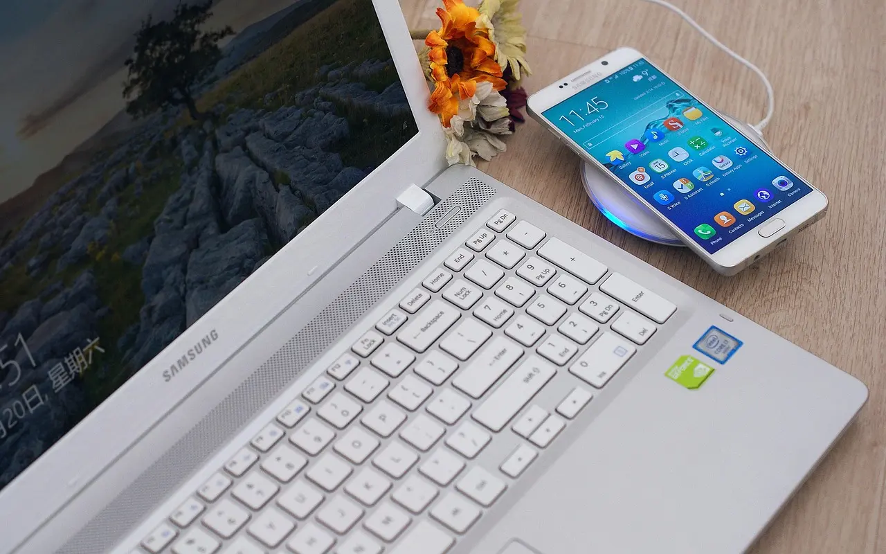 A Samsung Android phone showing its home screen on a wireless charger beside a laptop