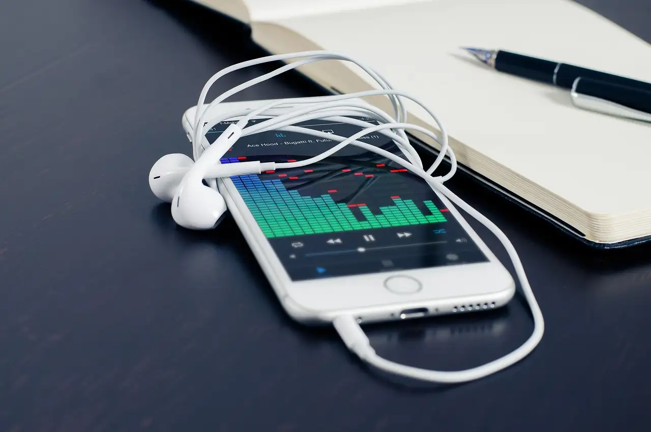 White smartphone on a dark desk shows a music equalizer, wired earbuds tangled across its screen