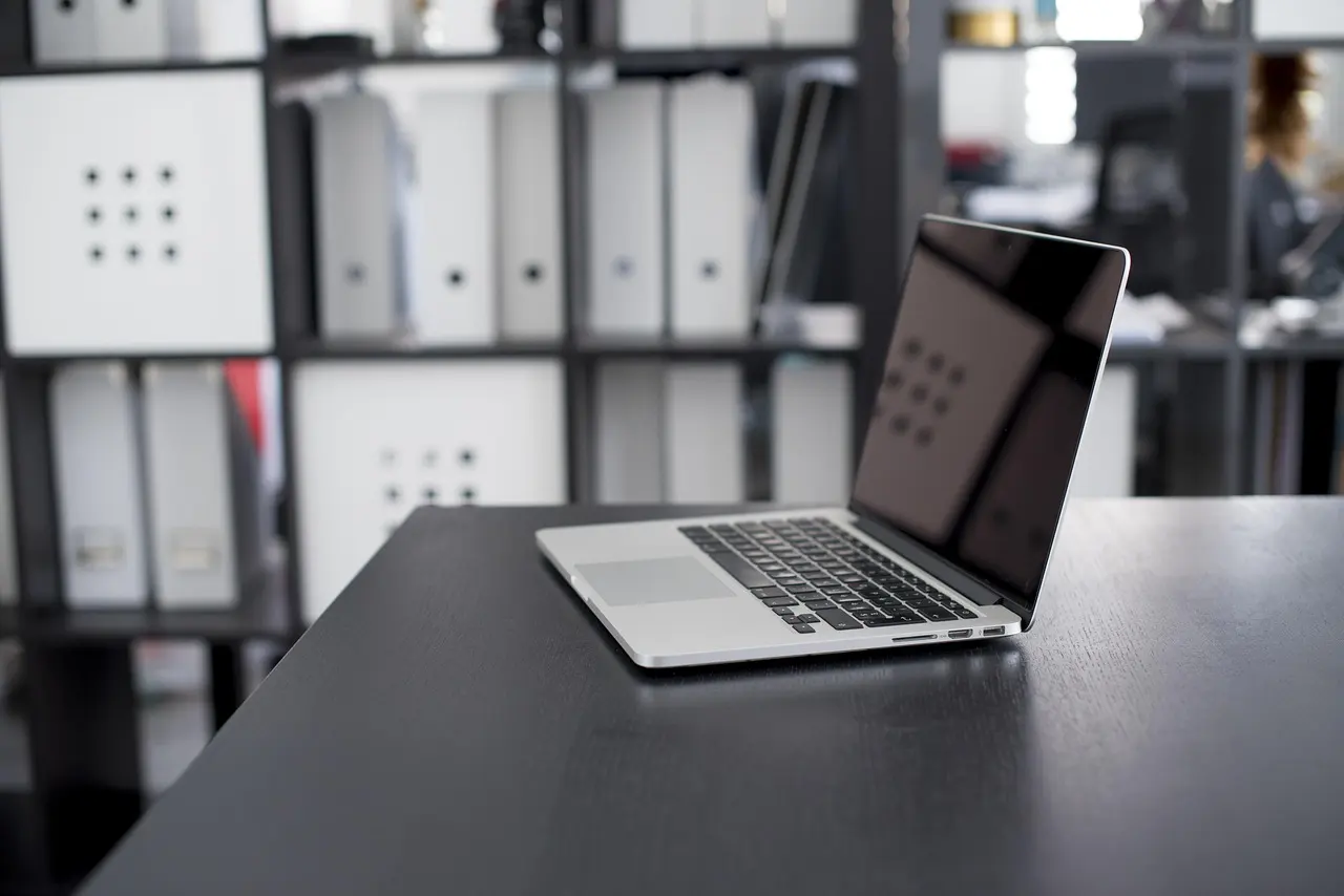 An open laptop with a blank screen on an office desk in front of shelves of binders