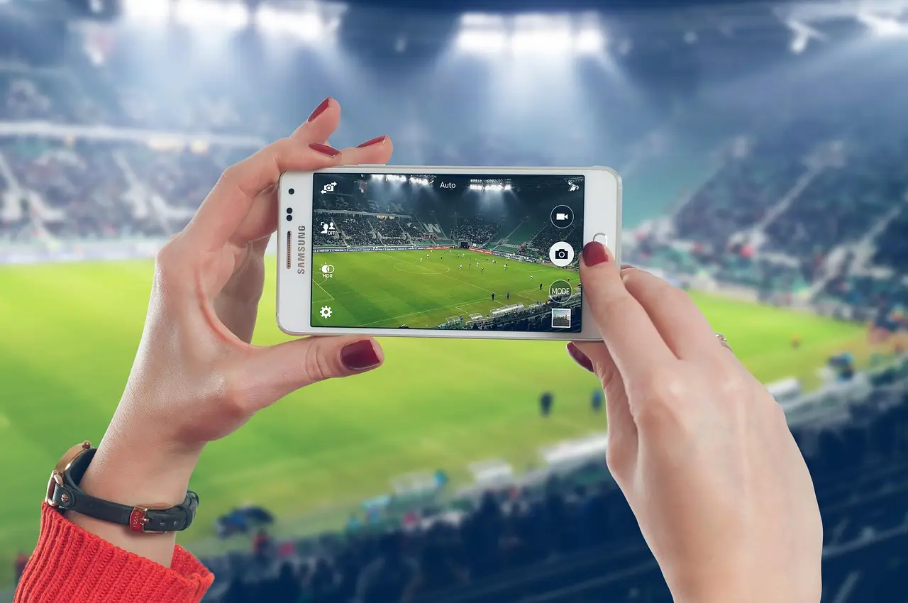Two hands holding a white Samsung phone with its camera aimed at a soccer stadium