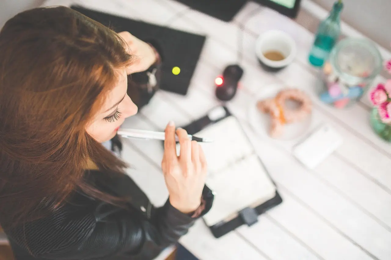 Overhead view of a woman writing in a planner with a pen beside coffee on a white table