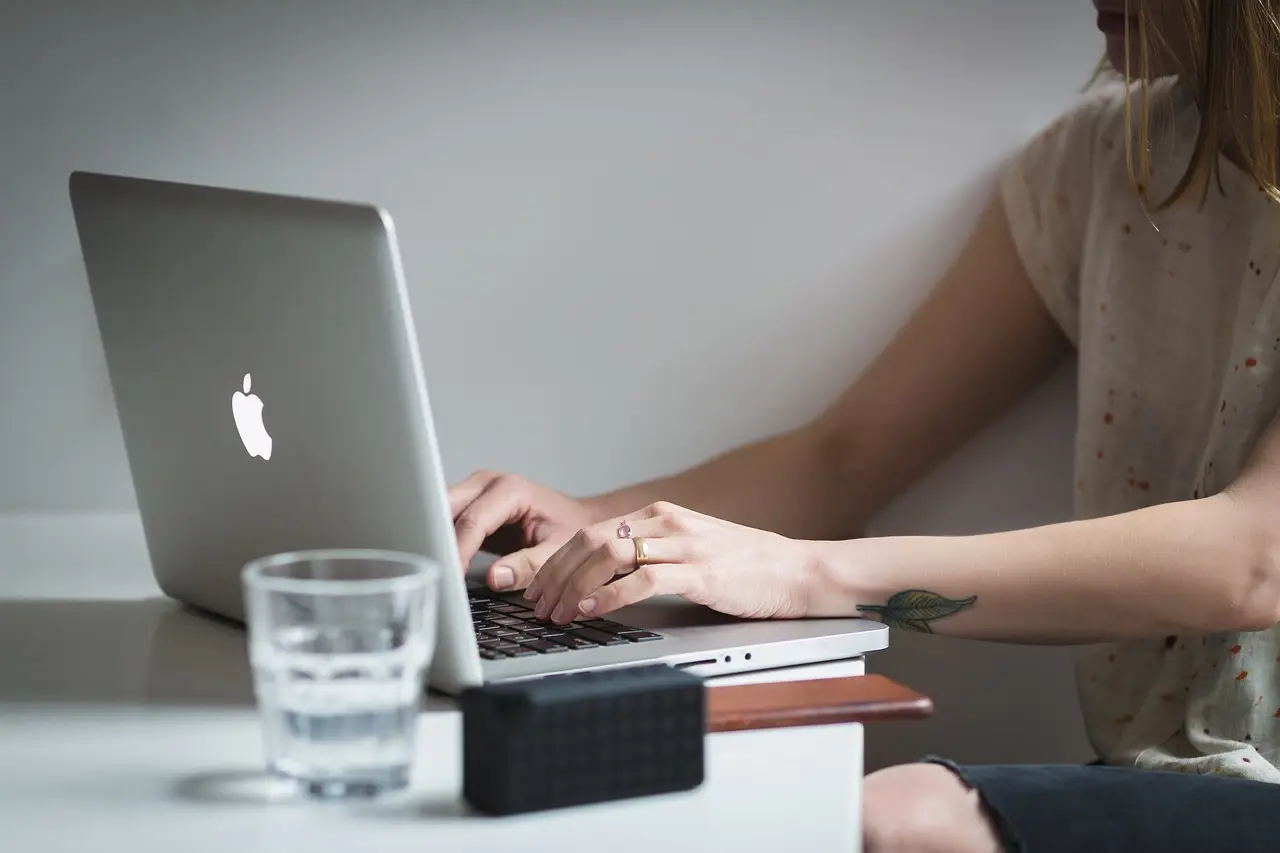 A woman typing on a laptop at a desk with a glass of water and a small speaker