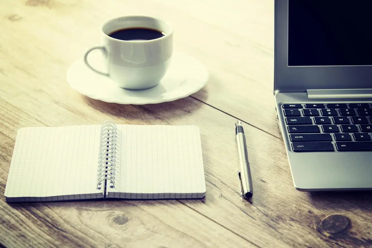 A cup of black coffee, an open spiral notebook with a pen, and a laptop on a wooden desk