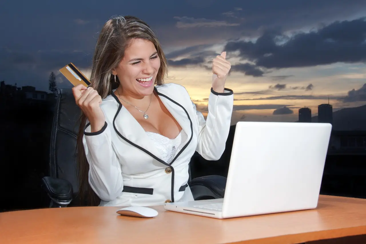 Woman in a white blazer cheers with a gold credit card beside a white laptop at dusk