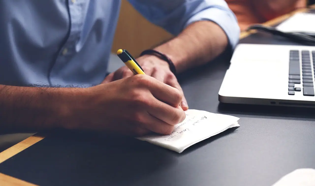 A person taking notes at a desk with a laptop.