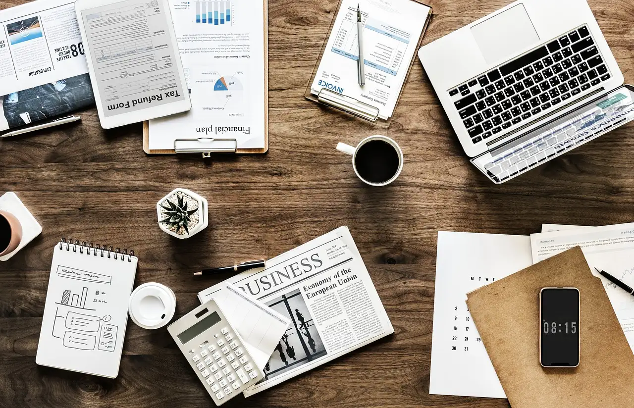 Overhead view of a wooden desk crowded with a laptop, newspaper, calculator, coffee, tax forms, and a phone