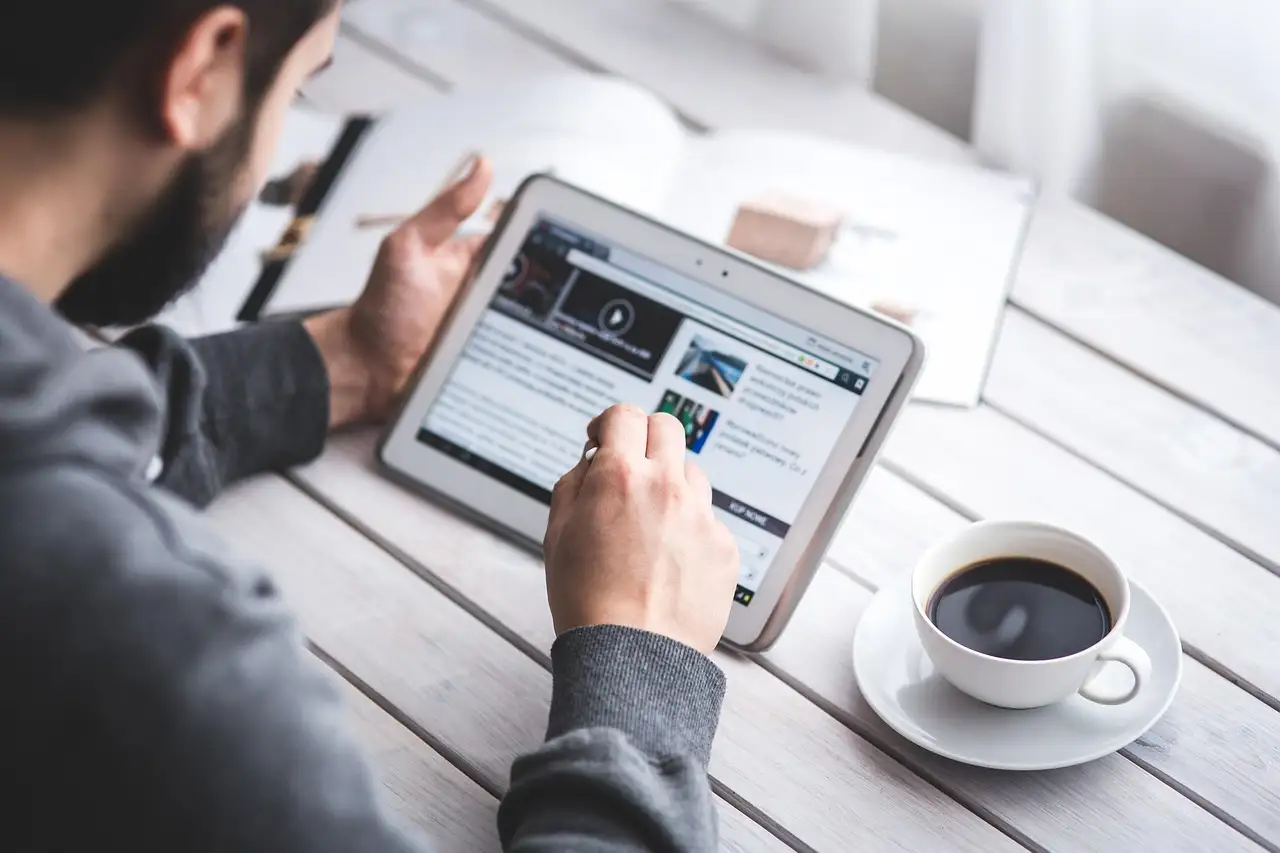 Bearded man in a gray hoodie taps a white tablet held sideways beside a cup of black coffee