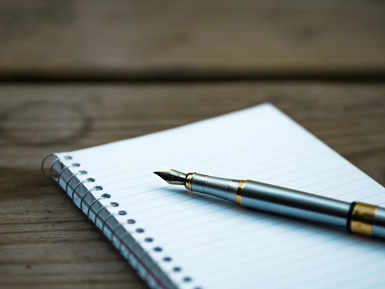 A fountain pen resting on a blank lined spiral notepad on a wooden table