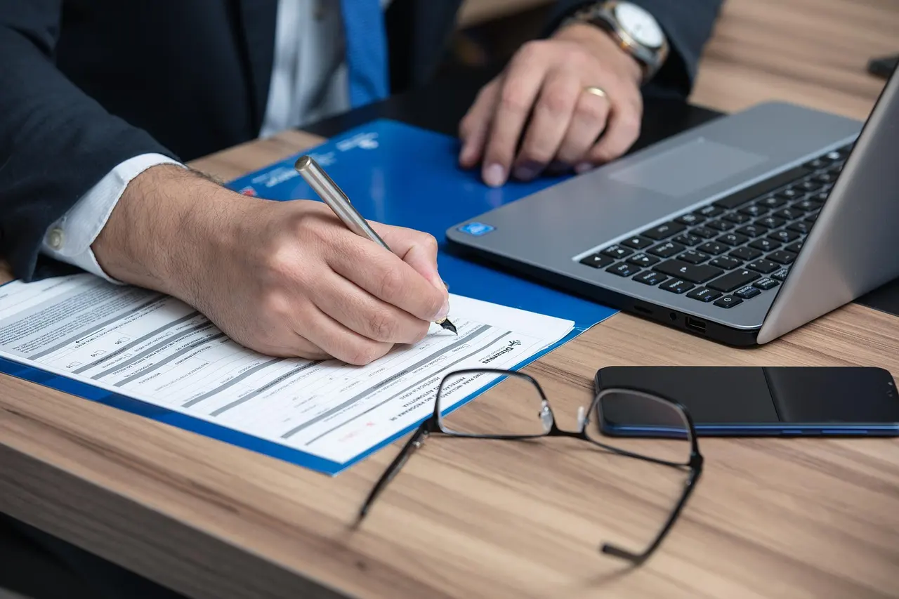 A person in a suit filling out a printed form with a pen beside a laptop and glasses on a desk