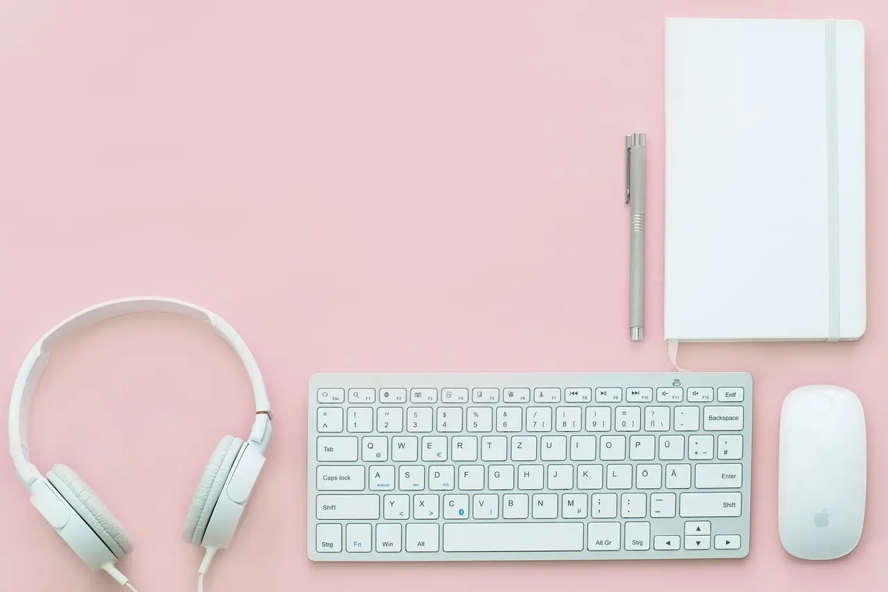A white keyboard, mouse, headphones, and a notebook arranged on a pink surface