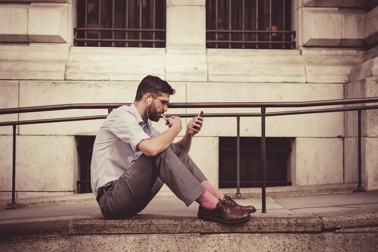 A man in a shirt sits on stone steps outdoors with earphones in, looking at the phone held in his hand