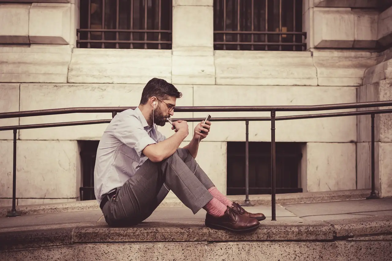 A man in a shirt sits on stone steps outdoors with earphones in, looking at the phone held in his hand
