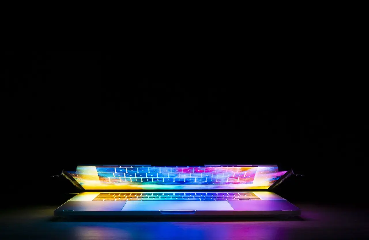 Open laptop in a dark room, screen and backlit keyboard glowing in rainbow colors