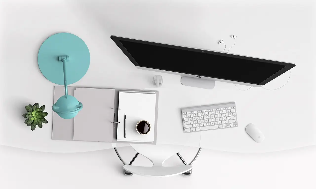 Overhead view of a computer monitor, keyboard, mouse, and binder on a white desk