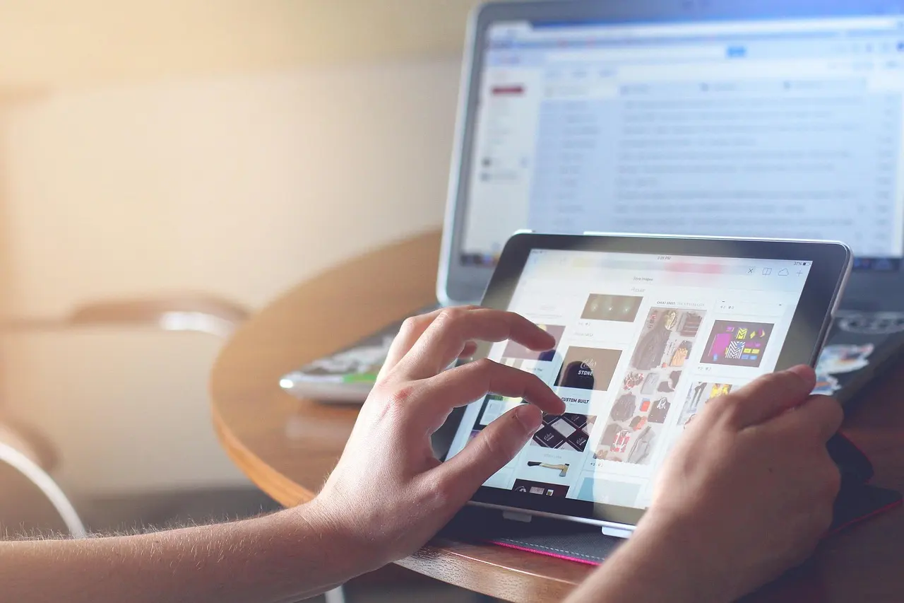 Hands using a tablet at a table, an open email inbox visible on a laptop screen behind it