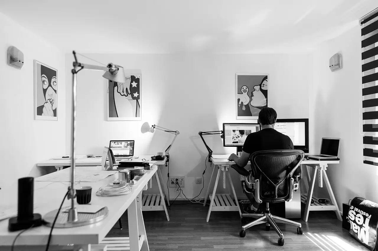 Hands writing in an open notebook on a wooden desk beside a MacBook Air and a phone showing a photo feed