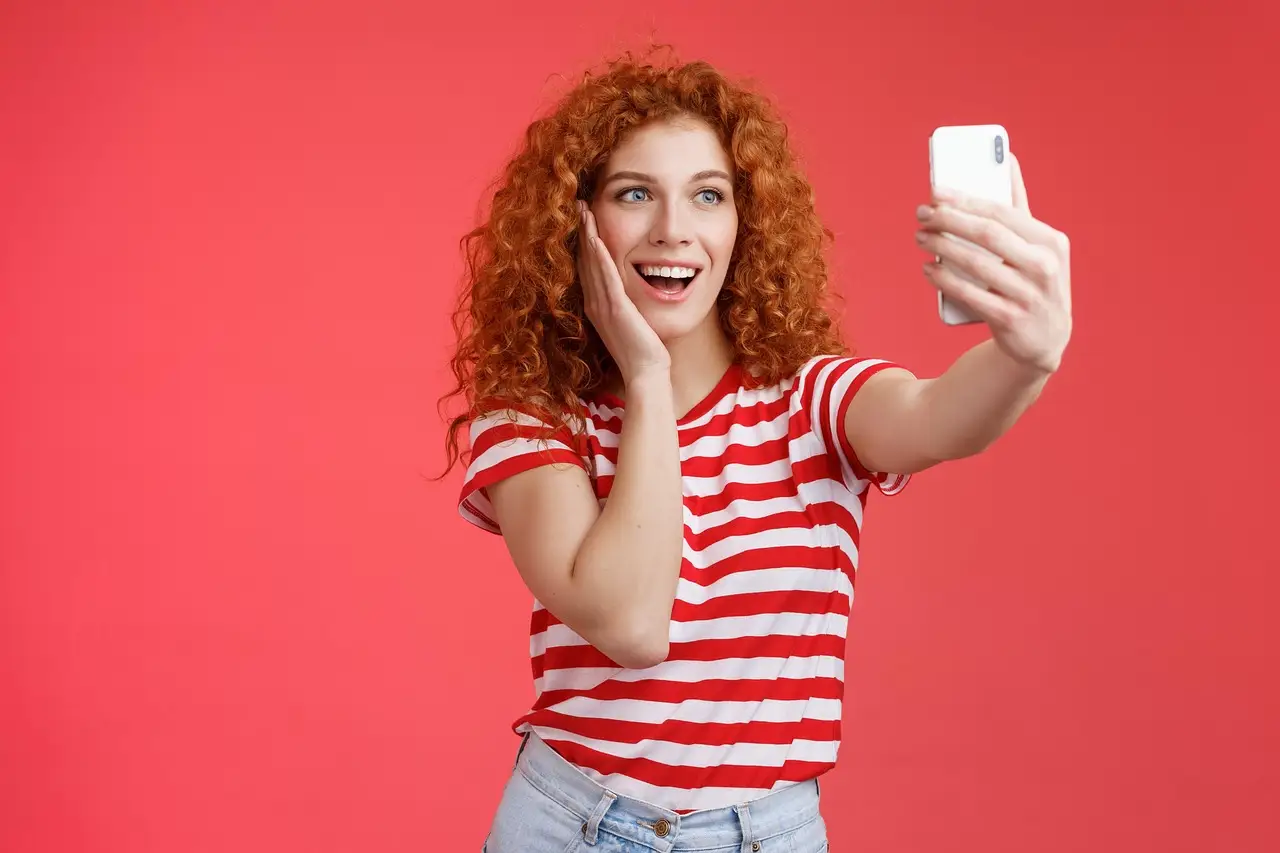 Curly red-haired woman in a striped tee holds a white smartphone at arm's length against a red backdrop