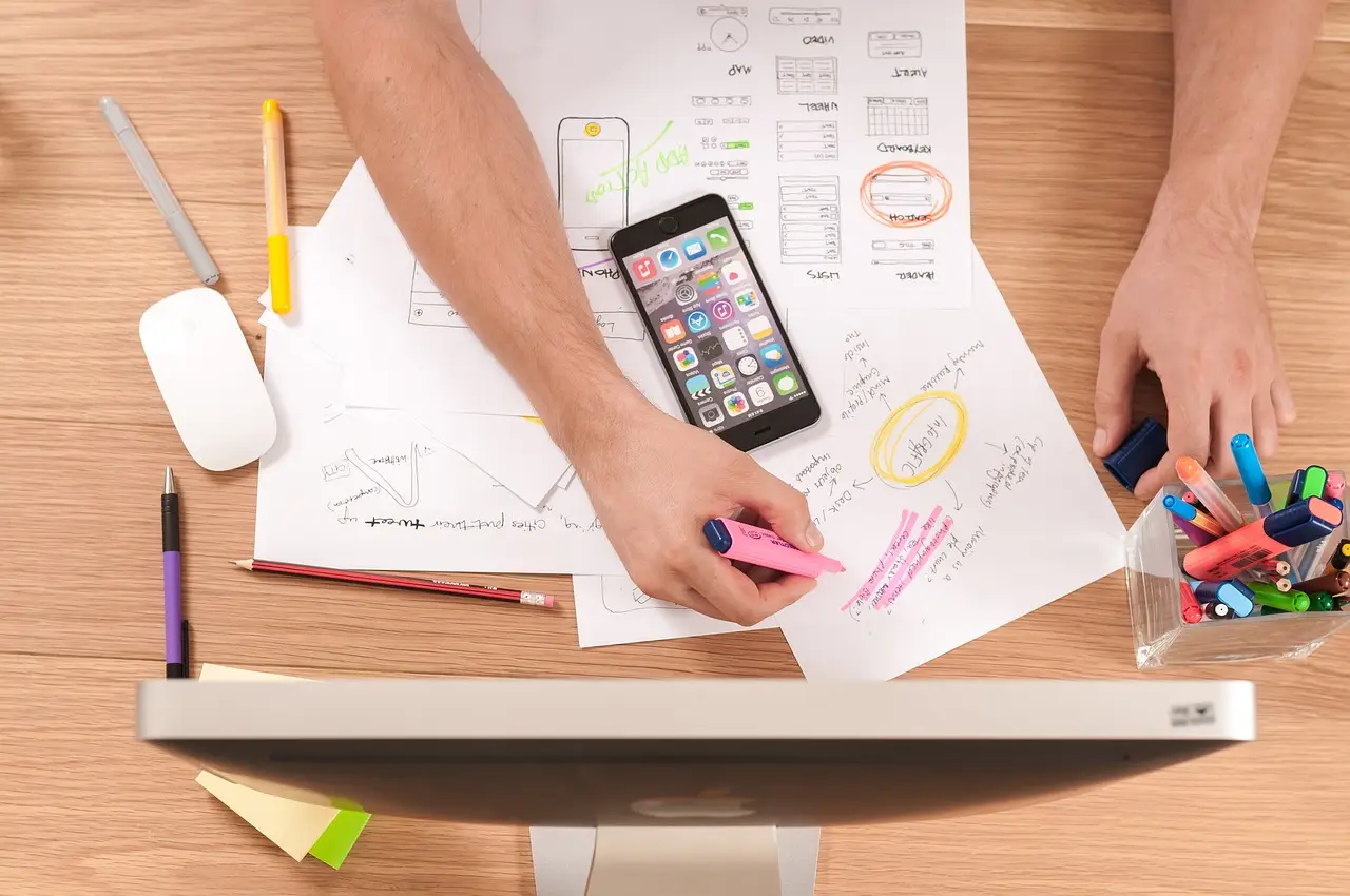 Overhead view of hands marking app wireframe sketches with a pink highlighter beside a phone and markers