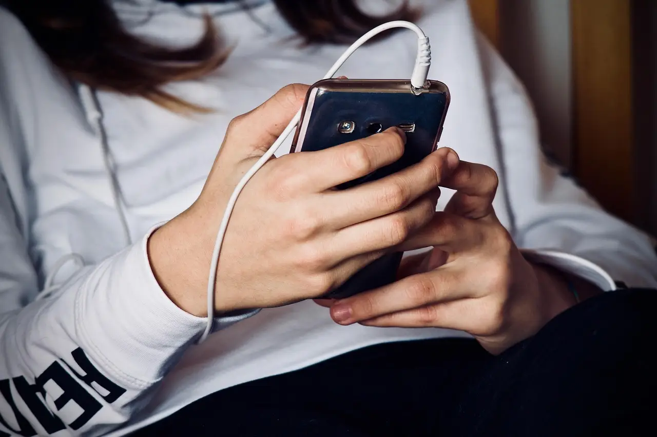 Close-up of hands holding a dark smartphone with a white wired earbud cable plugged into the top