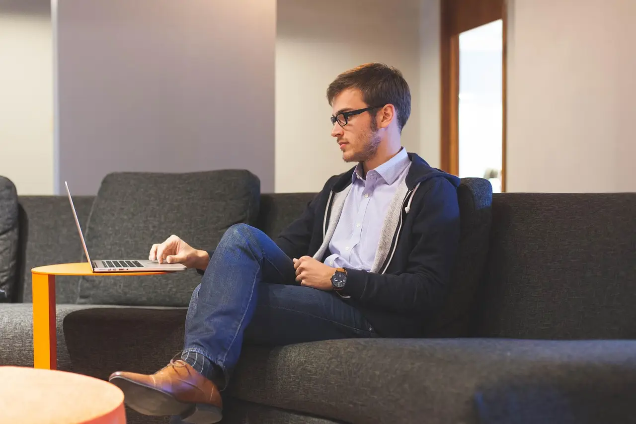 Man in glasses and a hoodie types on a laptop resting on an orange table beside a gray couch