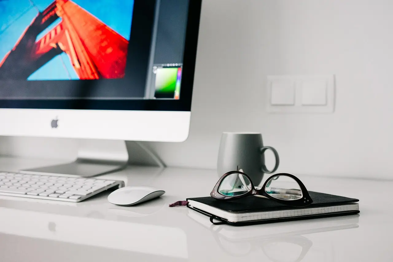 An iMac, wireless keyboard and mouse, and glasses on a notebook beside a mug on a white desk