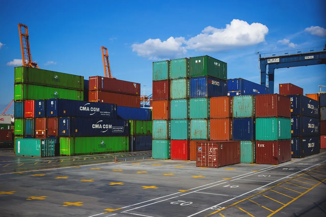 Stacks of green, blue, and rust shipping containers in a port yard under blue sky, cranes behind
