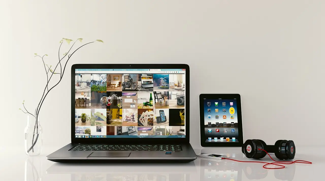 Hands tap a tablet showing a grid of product thumbnails, an open laptop behind on a wooden table